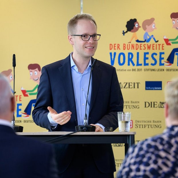 Dr. Jens Brandenburg, ehem. Parlamentarischer Staatssekretär bei der Bundesministerin für Bildung und Forschung, spricht bei einer Pressekonferenz zum Bundesweiten Vorlesetag 2023 an einem Rednerpult mit Mikrofon. Hinter ihm eine gelbe Pressewand mit dem Logo des Vorlesetags und den Logos der Initiatoren DIE ZEIT, Stiftung Lesen und Deutsche Bahn Stiftung. Im Vordergrund sitzen Dr. Jörg F. Maas Hauptgeschäftsführer der Stiftung Lesen und Sandra Kreft, Mitglied der Geschäftsleitung der ZEIT Verlagsgruppe. Die Szene vermittelt einen offiziellen Auftritt im Rahmen der Leseförderung. © Phil Dera