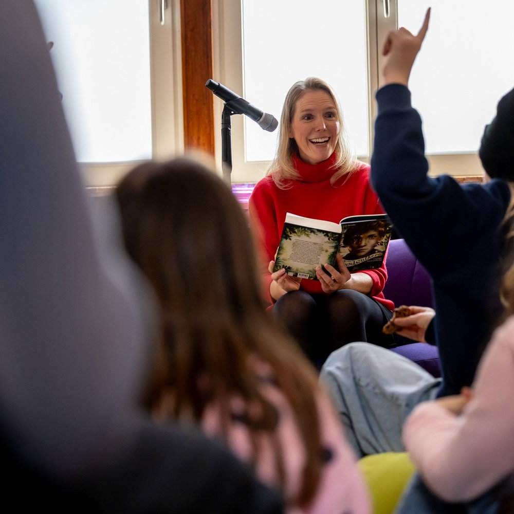 In einem hellen Raum sitzt Lesebotschafterin Maja Sommer in einem roten Pullover auf einem Stuhl und liest einer Gruppe von Kindern aus einem aufgeschlagenen Buch vor. Sie lächelt die Kinder an, während eines von ihnen mit erhobener Hand eine Frage stellen möchte. Hinter der Frau steht ein Mikrofon, die Kinder sitzen auf dem Boden und hören aufmerksam zu.