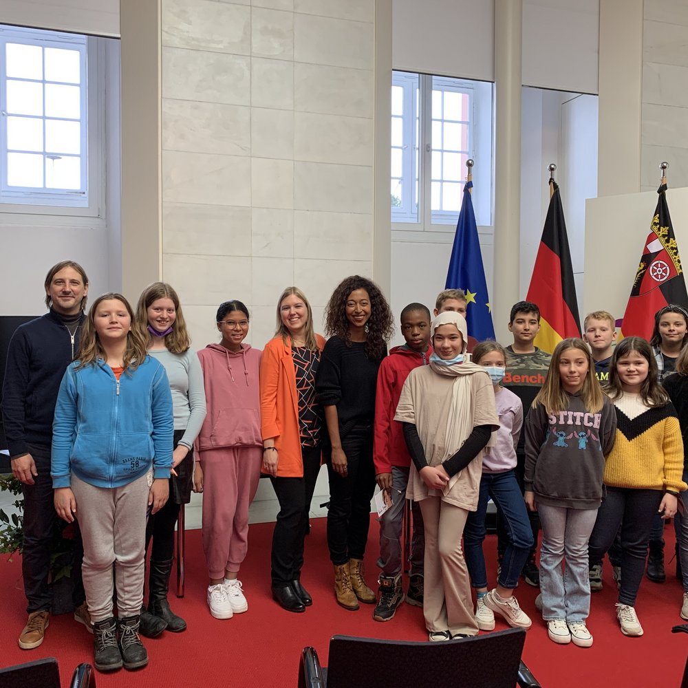 Gruppenfoto mit Jugendlichen und Erwachsenen in einem offiziellen Saal mit rotem Teppich. Im Hintergrund stehen die Flaggen der Europäischen Union, Deutschlands und von Rheinland-Pfalz. Die Gruppe besteht aus etwa 16 Personen, darunter Kinder, Jugendliche und vier Erwachsene darunter Melanie Raabe, Bestsellerautorin und Lesebotschafterin. Alle lächeln in die Kamera. Die Szene vermittelt Beteiligung, kulturelle Vielfalt und ein Bildungs- oder Vorleseformat im politischen Kontext.