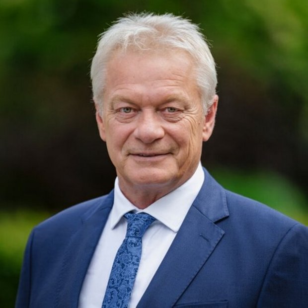 Ein Portrait von Bundesminister Alois Rainer, er trägt einen dunkelblauen Anzug, ein weißes Hemd und eine gemusterte blaue Krawatte. Er steht im Freien vor unscharfem, grünem Hintergrund und blickt freundlich in die Kamera.