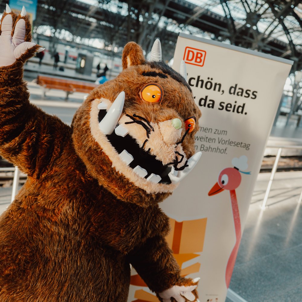 Der Grüffelo winkt fröhlich in die Kamera. Im Hintergrund steht ein Roll-Up der Deutschen Bahn mit der Aufschrift „Schön, dass ihr da seid.“