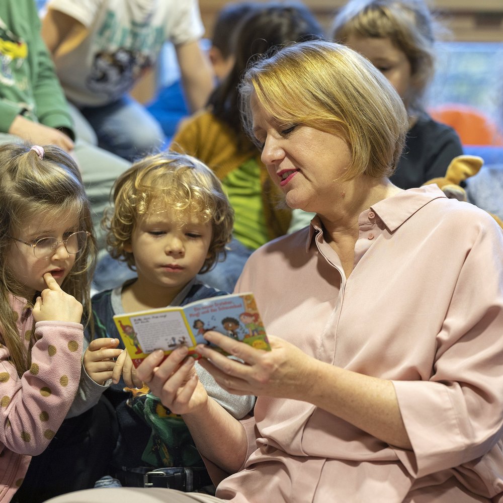 Lisa Paus, Bundesministerin für Familie, Senioren, Frauen und Jugend, sitzt mit mehreren Kita-Kindern auf einem blauen Sofa und liest aus einem kleinen Bilderbuch vor. Zwei Kinder schauen besonders konzentriert auf die Buchseiten, eines davon lutscht am Daumen. Weitere Kinder sitzen im Hintergrund. Die Szene vermittelt Nähe, Geborgenheit und Freude am gemeinsamen Vorlesen im Kita-Alltag.