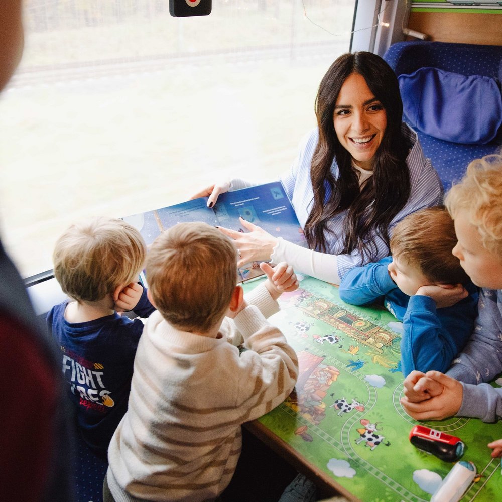 Moderatorin und Lesebotschafterin Jess Schöne sitzt im ICE in einer Ecke mit mehreren Sitzen. Um sie herum sind Kinder, denen sie vorliest. Dabei zeigt sie den umsitzenden Kindern die Innenseite des Buches, damit alle die Illustrationen sehnen können. Alle wirken fröhlich und entspannt, die Kinder schauen gebannt in das Buch. Insgesamt vermittelt das Bild eine fröhliche Atmosphäre.