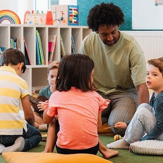 Ein pädagogischer Fachkraft liest sechs Kindern im Kita-Alter in einer hellen, gemütlich eingerichteten Gruppe vor. Die Kinder sitzen im Kreis auf Kissen auf einem grünen Teppich und hören gespannt zu.