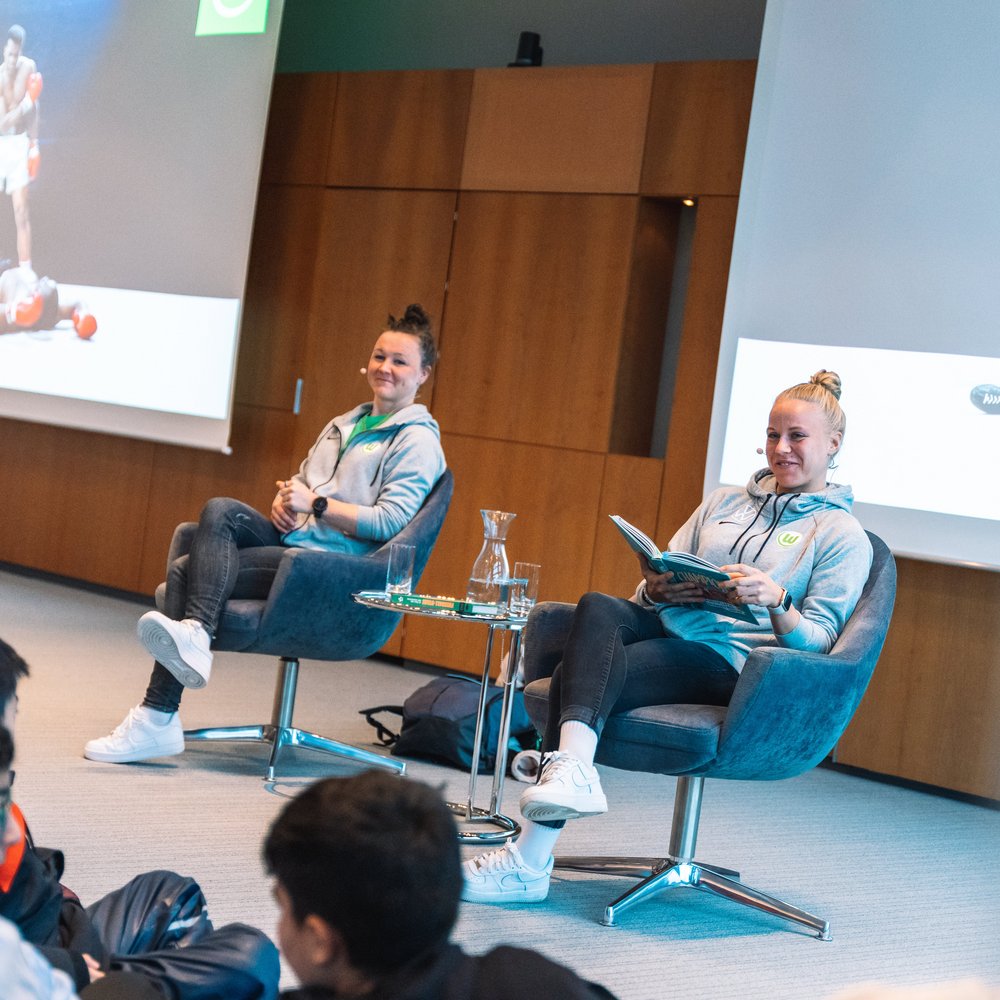 Marina Hegering und Pia-Sophie Wolter, Vfl Wolfsburg, sitzen in sportlicher Kleidung auf Sesseln in einem modernen Veranstaltungsraum und lesen Jugendlichen aus einem Buch vor. Im Hintergrund sind zwei große Leinwände zu sehen, die das ikonische Box-Foto von Muhammad Ali zeigen. Vor den Frauen sitzen mehrere Schüler und hören aufmerksam zu. Die Szene verbindet Vorlesen mit sportlichem Vorbildcharakter in einer motivierenden Lernumgebung.