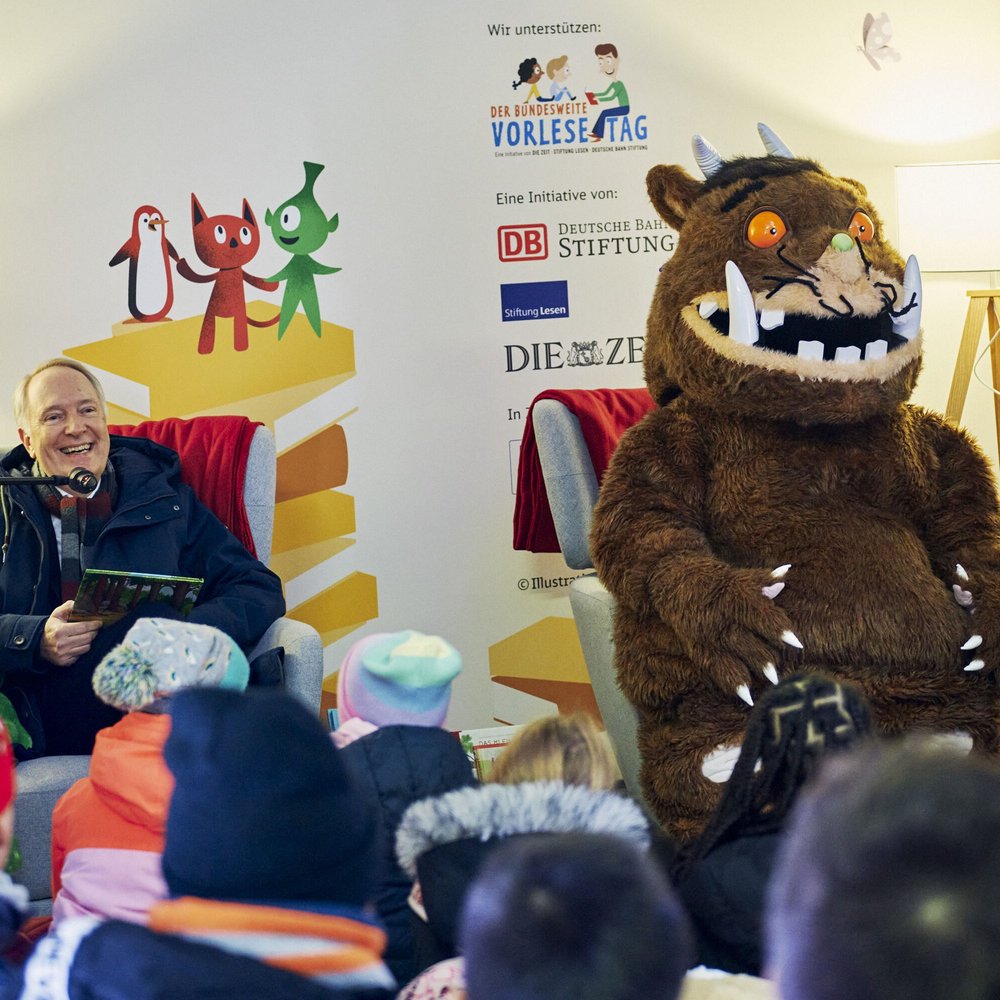Alexander Kaczmarek sitzt lächelnd in einem Sessel und liest Kindern aus einem Buch vor. Neben ihm sitzt der Grüffelo mit orangefarbenen Augen und langen Zähnen. Vor der Bühne sitzen viele Kinder in bunten Winterjacken und Mützen und hören gespannt zu. Im Hintergrund sind Illustrationen, Logos des Bundesweiten Vorlesetags sowie eine Stehlampe zu sehen.