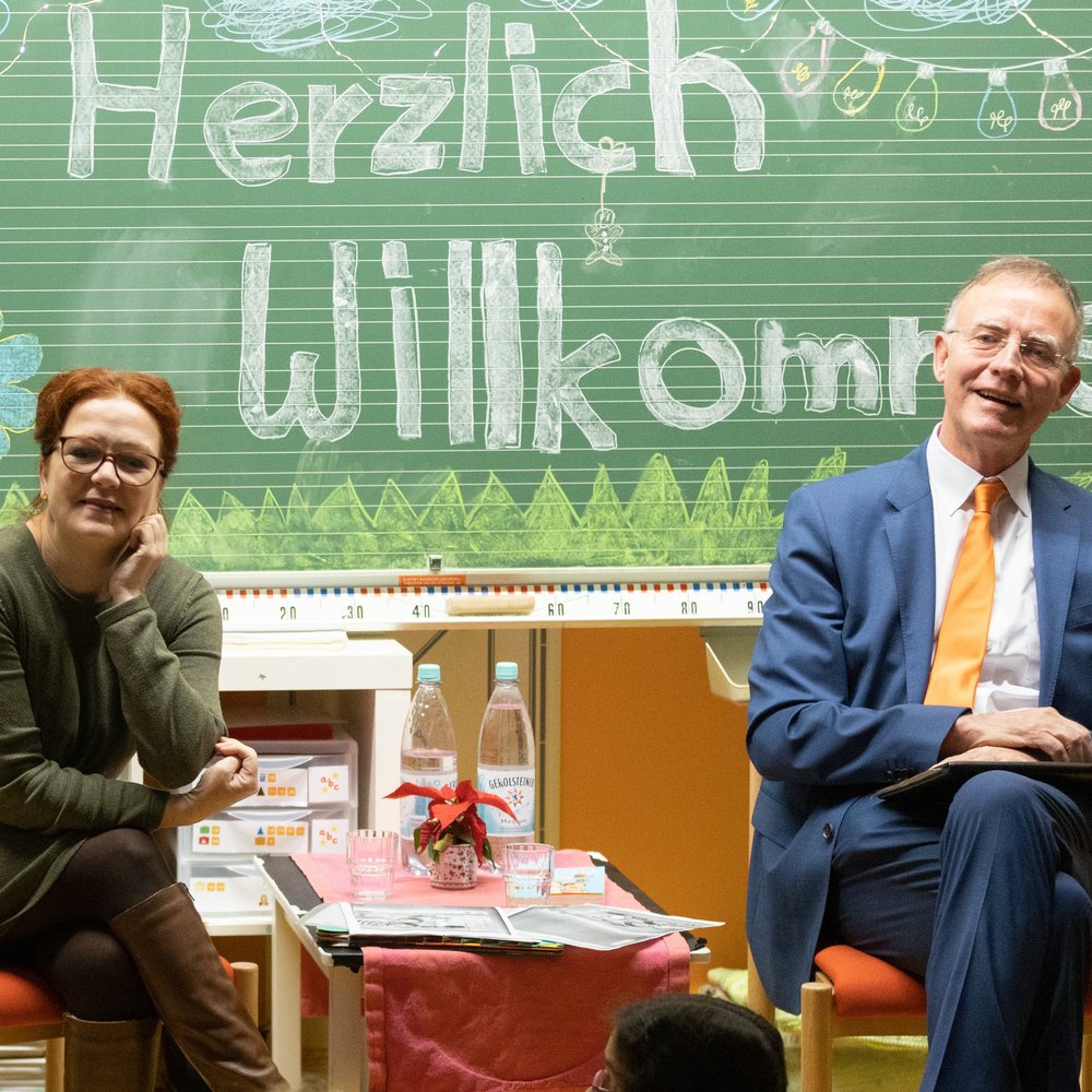Katja Dörner, Oberbürgermeisterin von Bonn und Dr. Gerd Landsberg, Deutscher Städte- und Gemeindebund, sitzen in einem bunt dekorierten Klassenraum vor einer Tafel mit der Aufschrift „Herzlich Willkommen“. Katja Dörner links trägt ein grünes Kleid und Stiefel, Dr. Gerd Landsberg rechts einen blauen Anzug mit orangefarbener Krawatte. Zwischen ihnen steht ein kleiner Tisch mit Wasserflaschen und Unterlagen. Im Vordergrund sind Kinderköpfe zu sehen. Die Szene vermittelt eine freundliche Vorlesesituation oder Begrüßungsveranstaltung in der Schule.