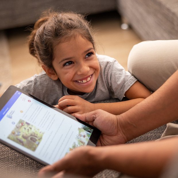 Lächelndes Mädchen liegt auf dem Sofa und schaut begeistert auf ein Tablet, das eine erwachsene Person in der Hand hält. Auf dem Bildschirm ist eine Vorlesegeschichte von „einfach vorlesen!“ zu sehen.