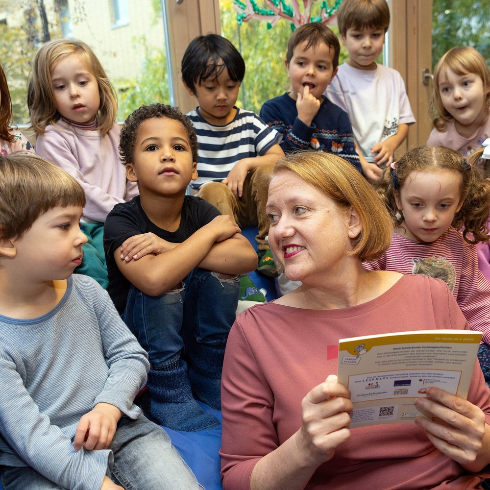 Lisa Paus, Bundesministerin für Familie, Senioren, Frauen und Jugend, liest einer großen Gruppe Kinder in gemütlicher Runde vor. Die Kinder sitzen dicht gedrängt um sie herum auf einem Sofa und hören gespannt zu – einige lächeln, andere schauen neugierig oder nachdenklich. Die Vorleserin hält ein illustriertes Kinderheft in der Hand. Im Hintergrund sind große Fenster mit Blick ins Grüne. Die Szene vermittelt Nähe, Lesefreude und frühkindliche Leseförderung.