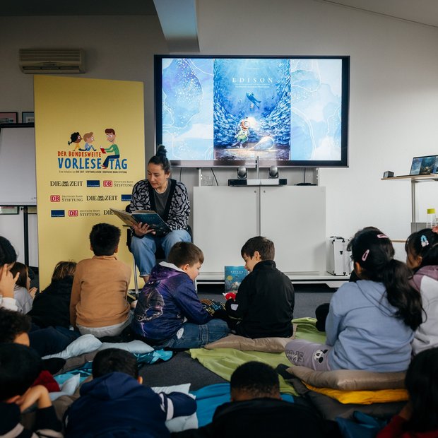 In einem Klassenraum liest eine Frau einer Gruppe von Kindern aus einem Bilderbuch vor. Die Kinder sitzen auf bunten Kissen am Boden. Neben der Frau steht ein gelbes Roll-Up mit dem Logo des Bundesweiten Vorlesetags. Im Hintergrund ist auf einem Bildschirm das Buchcover „Edison“ zu sehen.