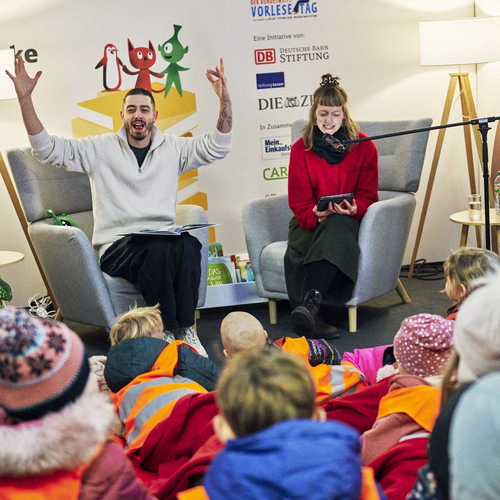Zwei Vorlesende sitzen in gemütlichen Sesseln auf einer kleinen Bühne. Jason Giuranna links hebt begeistert beide Arme, während er aus einem Buch vorliest. Die Frau rechts hält ein Tablet und lächelt. Vor der Bühne sitzen viele Kinder in bunten Jacken und orangefarbenen Warnwesten und hören aufmerksam zu. Im Hintergrund sind Figuren und Logos des Bundesweiten Vorlesetags sowie zwei Stehlampen und Pflanzen zu sehen.