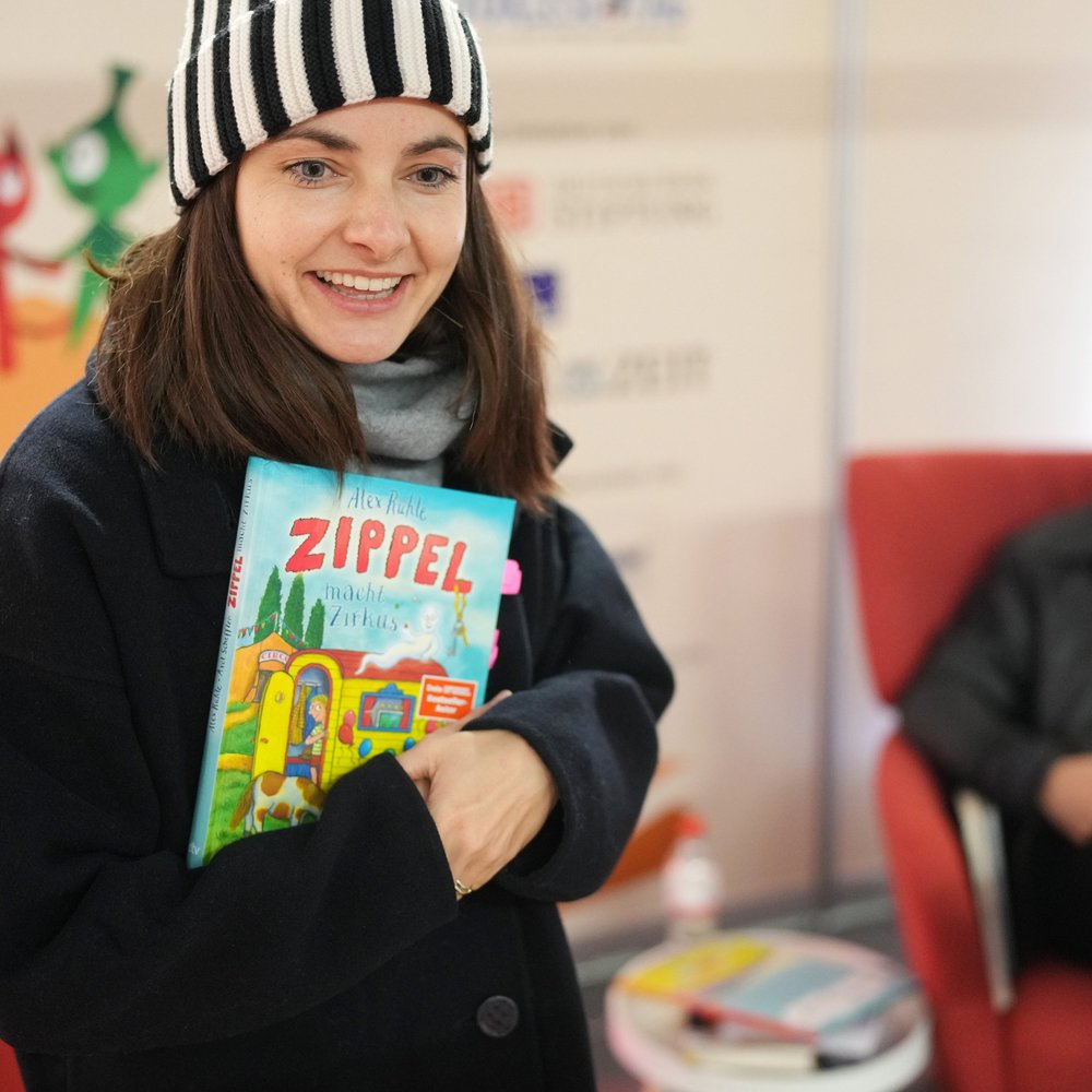 Paula Schramm, Lesebotschafterin und Moderatorin, steht im dicken Wintermantel mit Wollmütze im Vorlesebahnhof der DB. In der Hand hält sie ein Vorlesebuch, das sie gleich vorlesen wird. im Hintergrund ist es fröhlich dekoriert, passend zum Bundesweiten Vorlesetag. Laurence Rupp sitzt hinter ihr auf einem roten Sessel.  copyright: Deutsche Bahn Stiftung / Oliver Lang