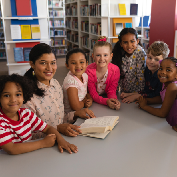 Eine junge Frau sitzt mit sechs Kindern unterschiedlicher Herkunft an einem Tisch in einer Bibliothek. Alle lächeln in die Kamera. Vor ihnen liegt ein aufgeschlagenes Buch.