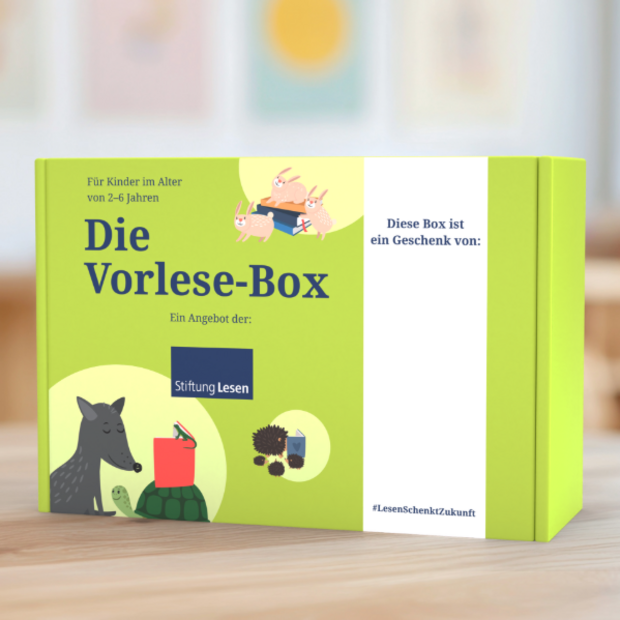 Grün gestaltete Vorlese-Box mit der Aufschrift „Die Vorlese-Box – Ein Angebot der Stiftung Lesen“. Oben steht: „Für Kinder im Alter von 2–6 Jahren“. Die linke Seite zeigt Illustrationen von Tieren wie Wolf, Igel, Hase und Schildkröte mit Büchern. Rechts befindet sich ein weißes Feld mit dem Text „Diese Box ist ein Geschenk von:“ sowie der Hashtag #LesenSchenktZukunft. Die Box steht auf einem Holztisch vor verschwommenem Hintergrund.